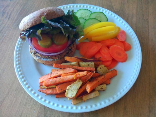 Black bean burger with red onion, tomato, pickles, and lettuce on a bun with a side of sweet potato and yellow squash fries, and raw slices of carrots, cucumbers, and yellow bell pepper